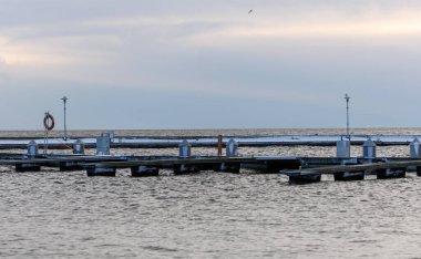 Nida Limanı 'nda kışın boş bir marina. Buzla kaplı yüzen iskeleler, sakin sular, bulutlu gökyüzü ve kıyı manzarası. İnsan yok, tekne yok. Soğuk mevsim, sessizlik, asgari deniz geçmişi.