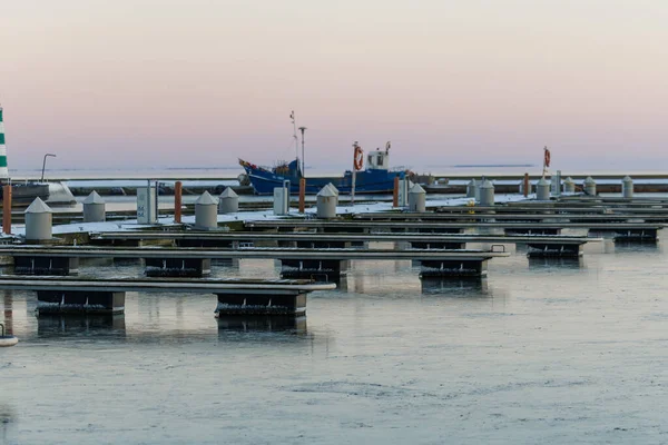 Nida Limanı 'nda kışın boş bir marina. Buzla kaplı yüzen iskeleler, sakin sular, bulutlu gökyüzü ve kıyı manzarası. İnsan yok, tekne yok. Soğuk mevsim, sessizlik, asgari deniz geçmişi.