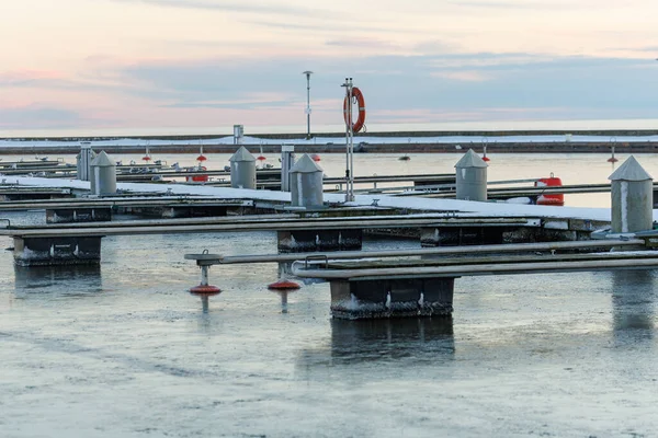 Nida Limanı 'nda kışın boş bir marina. Buzla kaplı yüzen iskeleler, sakin sular, bulutlu gökyüzü ve kıyı manzarası. İnsan yok, tekne yok. Soğuk mevsim, sessizlik, asgari deniz geçmişi.