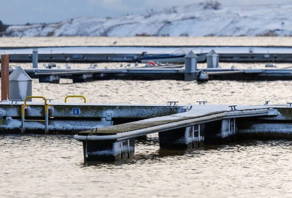Nida Limanı 'nda kışın boş bir marina. Buzla kaplı yüzen iskeleler, sakin sular, bulutlu gökyüzü ve kıyı manzarası. İnsan yok, tekne yok. Soğuk mevsim, sessizlik, asgari deniz geçmişi.