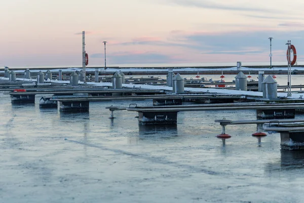 Nida Limanı 'nda kışın boş bir marina. Buzla kaplı yüzen iskeleler, sakin sular, bulutlu gökyüzü ve kıyı manzarası. İnsan yok, tekne yok. Soğuk mevsim, sessizlik, asgari deniz geçmişi.