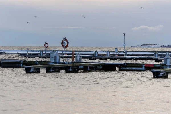 Nida Limanı 'nda kışın boş bir marina. Buzla kaplı yüzen iskeleler, sakin sular, bulutlu gökyüzü ve kıyı manzarası. İnsan yok, tekne yok. Soğuk mevsim, sessizlik, asgari deniz geçmişi.