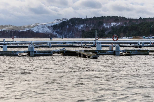 Nida Limanı 'nda kışın boş bir marina. Buzla kaplı yüzen iskeleler, sakin sular, bulutlu gökyüzü ve kıyı manzarası. İnsan yok, tekne yok. Soğuk mevsim, sessizlik, asgari deniz geçmişi.