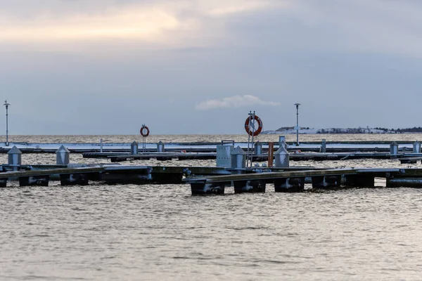 Nida Limanı 'nda kışın boş bir marina. Buzla kaplı yüzen iskeleler, sakin sular, bulutlu gökyüzü ve kıyı manzarası. İnsan yok, tekne yok. Soğuk mevsim, sessizlik, asgari deniz geçmişi.