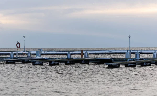 Nida Limanı 'nda kışın boş bir marina. Buzla kaplı yüzen iskeleler, sakin sular, bulutlu gökyüzü ve kıyı manzarası. İnsan yok, tekne yok. Soğuk mevsim, sessizlik, asgari deniz geçmişi.