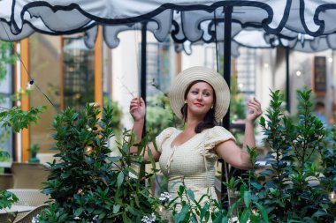 Açık sarı bir yaz elbisesi ve hasır şapka giyen zarif bir kadın yeşil bir kafeterya bahçesinde duruyor. Rahat şehir yaşam tarzı, kadınsı tarz, yaz tatili, sakin atmosfer, kopyalama alanı