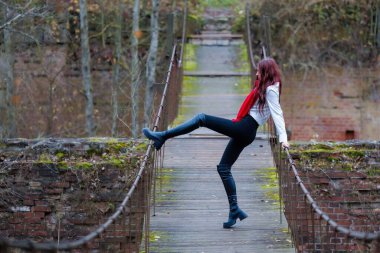 Kırmızı eşarp, beyaz gömlek ve siyah pantolon giyen genç bir kadın, sonbahar ormanıyla çevrili eski bir tahta köprü üzerinde etkileyici esneme ve dengeleyici pozlar veriyor. Kavramsal açık hava modası ve yaşam tarzı portresi hareketleri birleştiriyor, bal