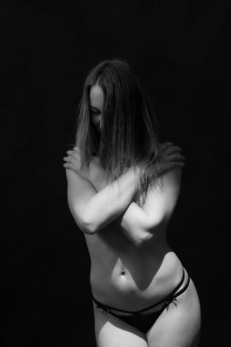 Yetişkin bir kadının kendini kucakladığı özel siyah beyaz bir stüdyo resmi. Kırılganlık, kendini kabullenme, kadınsılık ve iç gücü ifade eden sanatsal kompozisyon. En az koyu arkaplan.