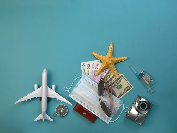 airplane model, passport with covered medical mask, ampoule with vaccine and syringe, dollar and euro bills, sunglasses, starfish and compass - what you need for a seaside holiday