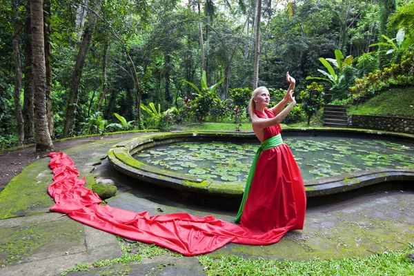 Beautiful girl in a red long dress standing in the enchanted forest near waterpool and resists wind — Stock Photo, Image