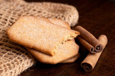 İki buçuk dikdörtgen şeker kaplı tarçınlı bisküvi. Yanında da tarçınlı çubuklar ve siyah ahşap masa..