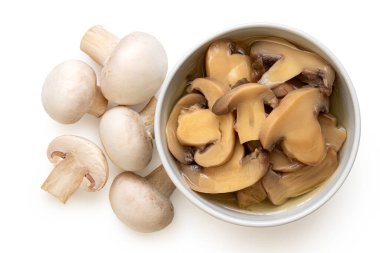 Dilimlenmiş mantar konservesi beyaz bir seramik kasenin içinde taze düğmeli mantarların yanında. Üst görünüm.