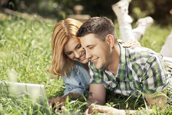 Glada unga par liggande i gräset, titta på laptop och smi Royaltyfria Stockbilder