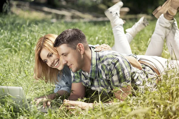 Glada unga par liggande i gräset, titta på laptop och smi Stockbild