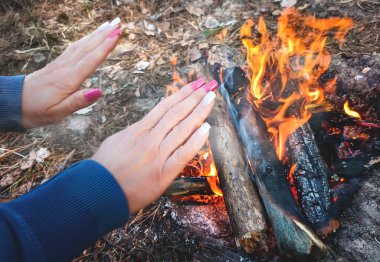 Kadın dışarıda, ormanda kamp yaparken ateş yakıyor. Ateşin yanında ellerini ısıtıyor.