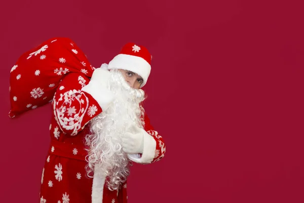 Papai Noel segura um saco de presentes atrás de suas costas em um fundo vermelho. — Fotografia de Stock