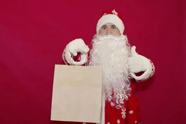 Санта тримає паперову сумку з подарунками і тримає пальці на червоному фоні.. — стокове фото
