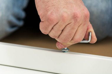 Bir adam, mavi gömlekli bir tamirci, bir masa topluyor, yakın plan. Diz çökerken masa çerçevesini uğursuz bir anahtarla birleştirir..