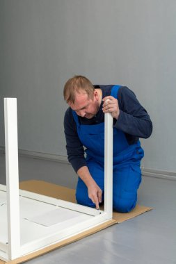Mavi tulumlu bir adam yakın plan bir masa kuruyor. Diz çökerken masa bacaklarını İngiliz anahtarıyla sabitler.. 