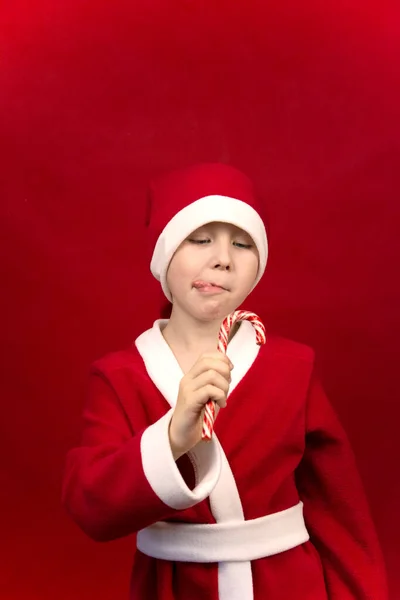 Um menino em um traje vermelho Papai Noel detém bengalas doces em suas mãos. — Fotografia de Stock