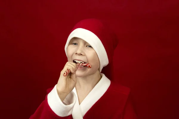 Um menino em um traje vermelho Papai Noel detém bengalas doces em suas mãos. — Fotografia de Stock