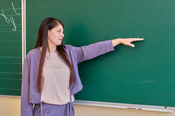 De jonge leraar wijst naar het schoolbord met haar hand, kopieer ruimte. — Stockfoto