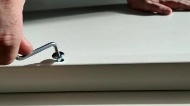 Bir adam, mavi gömlekli bir tamirci, bir masa topluyor, yakın plan. Diz çökerken masa çerçevesini uğursuz bir anahtarla birleştirir..