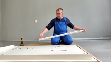 Bir adam, mavi tulumlu bir tamirci, yakın planda bir masa kuruyor. Diz çökerken masa çerçevesini elektrikli tornavidayla birleştiriyor..