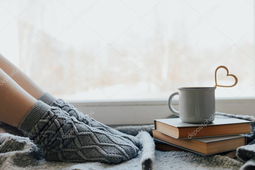 Cozy winter still life. Woman legs in warm woolen socks with mug of hot beverage on old windowsill against snow landscape. Love at distance, loneliness in self-isolation in the time of coronavirus.