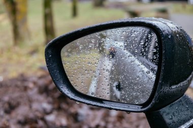 Close up of car side rear view mirror with rain drops on blurred background. Rainy day on the road. Driving in bad weather.