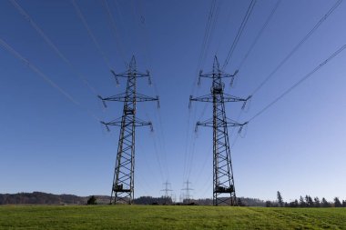 Sonbaharda yeşil bir çayırda parlak mavi bir gökyüzünün önünde iki büyük yüksek voltajlı elektrik direği, arka planda daha fazla elektrik direği görülebilir, modern toplumumuzda elektrik hayati önem taşır.