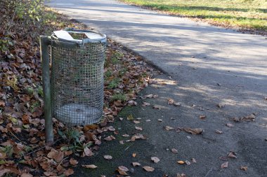 Ormanın gölgesinde boş kafes atığı kovası. Arka planda, gündüz vakti insanlar olmadan güneş ışığında gri asfaltı görebilirsiniz.