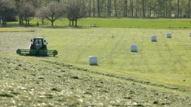 Büyük yeşil bir çayırda ilerleyen tarımsal makine samanlara işlenmiş çimenleri hasat ediyor. Bitmiş saman balyaları, gün boyunca, gün ışığında, tarlada beyaz poşetlere doldurulmuş durumda.