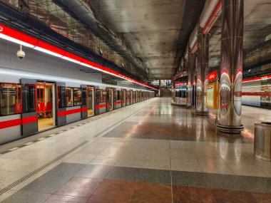 Prag, Çek Cumhuriyeti, 9 Mart 2026 Yolcular, bir metro treni modern yeraltı istasyonuna vardığında platformda yürümek ve beklemek, kıvrımlı tavanlar, parlak ışıklandırma, redandwhite tabelası ve günlük banliyö aktivitesi.