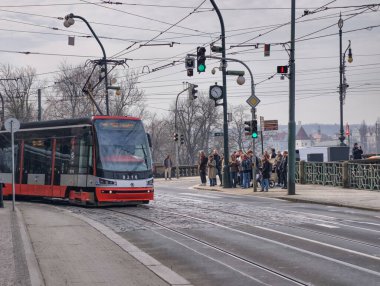 Prag, Çek Cumhuriyeti, 11 Mart 2026, Vltava nehri yakınlarındaki tarihi merkezde günlük yaşam ve ulaşım altyapısı. Yaya geçitlerinden geçen tramvaylar, yolcular ve yayaların yer aldığı şehrin gazetecilik manzarası.