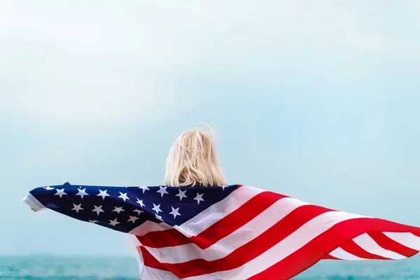 Biała blondynka trzymająca amerykańską flagę opierającą się o plecy i patrzącą na morze. Flaga USA na Dzień Niepodległości — Zdjęcie stockowe