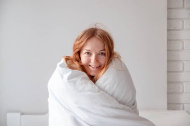 Gülen kız beyaz, sıcak bir battaniyeyle yatağın üzerinde oturuyor. Günaydın.