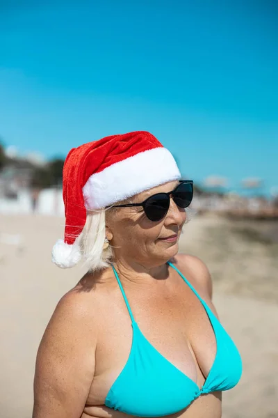 Retrato de mulher loira vestindo chapéu vermelho de Papai Noel comemorando as férias de Natal e sorrindo para a câmera junto ao mar em um dia ensolarado — Fotografia de Stock