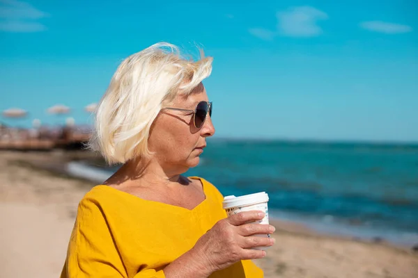 Vacker kvinna som håller papper kopp kaffe och njuter av en promenad längs havskusten — Stockfoto
