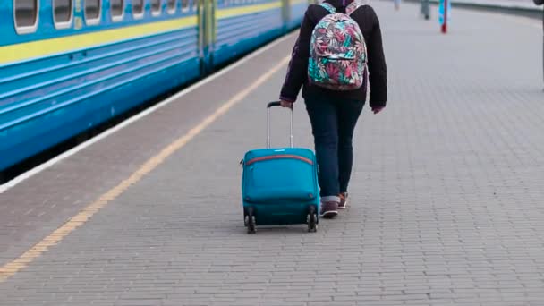 Kobieta z plecakiem i niebieską walizką spaceruje wzdłuż peronu do pociągu, widok z tyłu — Wideo stockowe
