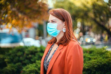 Tıbbi maskeli ve takım elbiseli genç bir kız elinde bir akıllı telefon tutuyor ve sokağa bakıyor. Coronavirus, covid 19 konsepti.