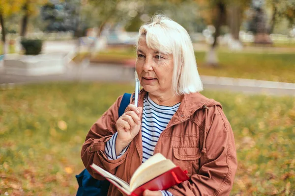 一个心事重重、忙碌的老妇人的画像，手里拿着记事本和钢笔，计划着，专心致志地分析着她在秋天的公园里散步。头脑风暴的概念 — 图库照片