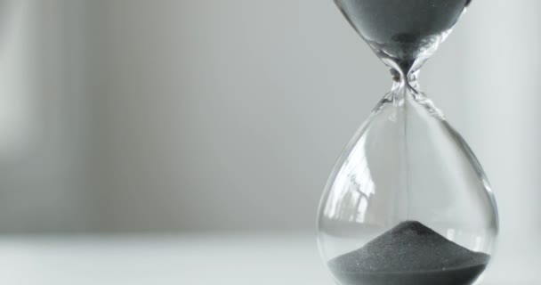 Durch die Sanduhr läuft grauer Sand. transparente Sanduhr aus Glas auf dem Hintergrund eines weißen Nachttisches. — Stockvideo