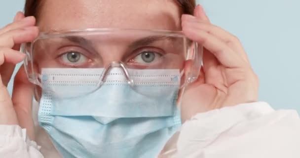 Close-up de um médico ou médico em equipamentos de proteção individual e luvas cirúrgicas azuis coloca óculos transparentes para proteger contra vírus, bactérias durante uma pandemia — Vídeo de Stock