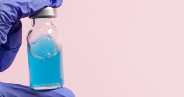 Médico o enfermero manos en guantes azules sostiene un frasco de vidrio con líquido de vacuna azul sobre un fondo rosa. — Vídeo de stock