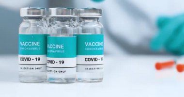 Aşı şişeleri beyaz bir laboratuvar masasında. Koruyucu üniformalı doktor elinde ilaç ve aşı şişesi tutuyor. Genel Aşılama.