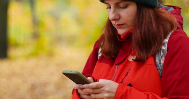 Chica joven en ropa deportiva utiliza un teléfono inteligente, escribe SMS en el parque. Viaje de temporada de otoño — Vídeo de stock