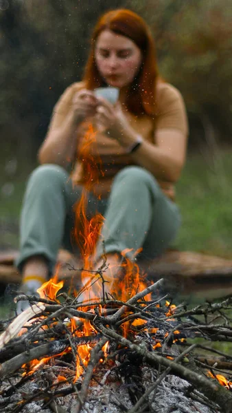 Arka planda sıcak bir içecek içen genç bir kız, sığ bir alan derinliği. Ormanda barbekü ya da bbq pikniği için şenlik ateşi — Stok fotoğraf