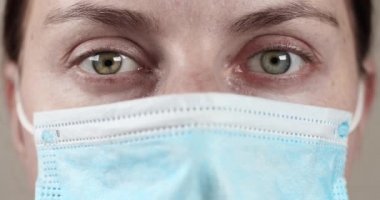 COVID-19 Sars-cov-2 salgını. Koruyucu tıbbi maskeli, gri gözlü genç bayan doktor kameraya bakıyor.