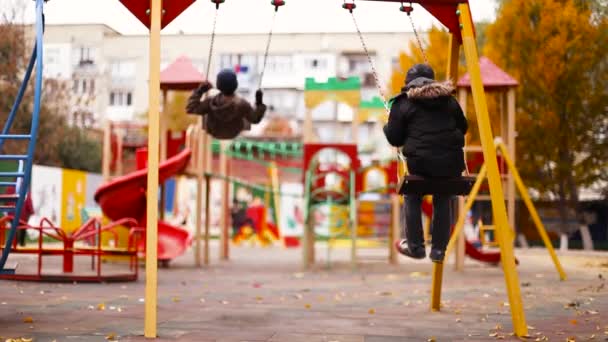 I bambini cavalcano un'altalena nel parco giochi. Ragazzi 8 10 anni in abiti caldi camminano nel parco della città — Video Stock